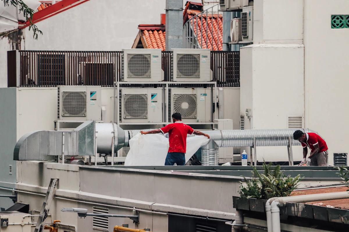 Two workers handling ductwork on a rooftop beside rows of outdoor air conditioning units