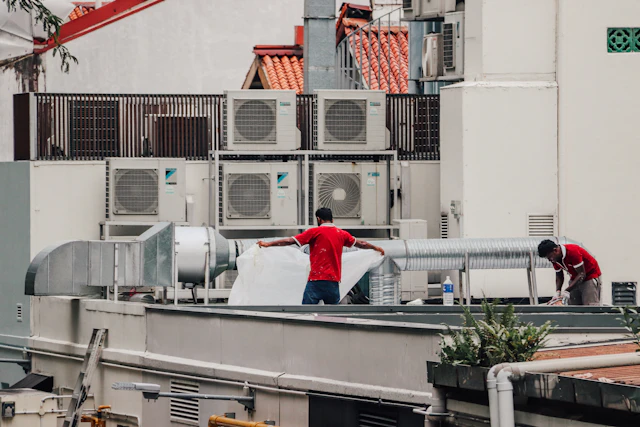 Two workers handling ductwork on a rooftop beside rows of outdoor air conditioning units